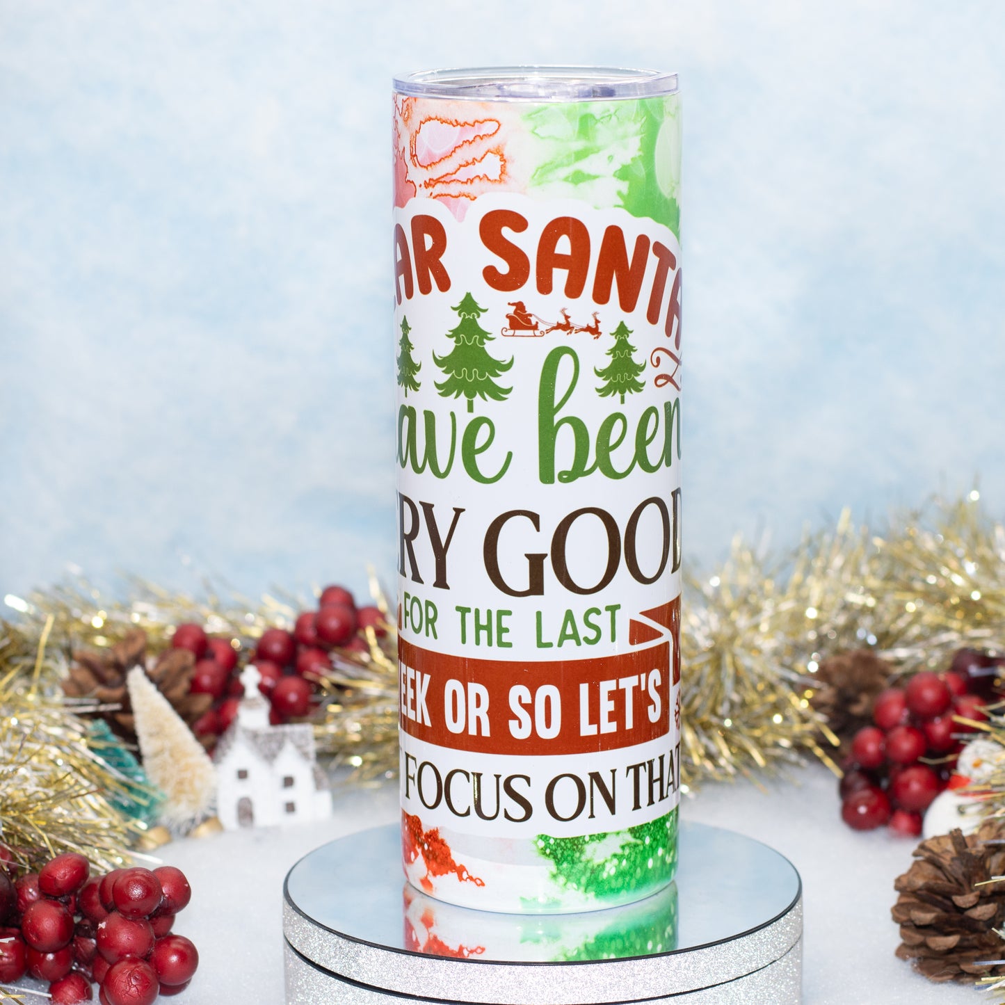An insulated tumbler with a colorful Christmas-themed graphic. The background is decorated with gold tinsel, red berries, pinecones, and a small white house against a soft light blue backdrop.
