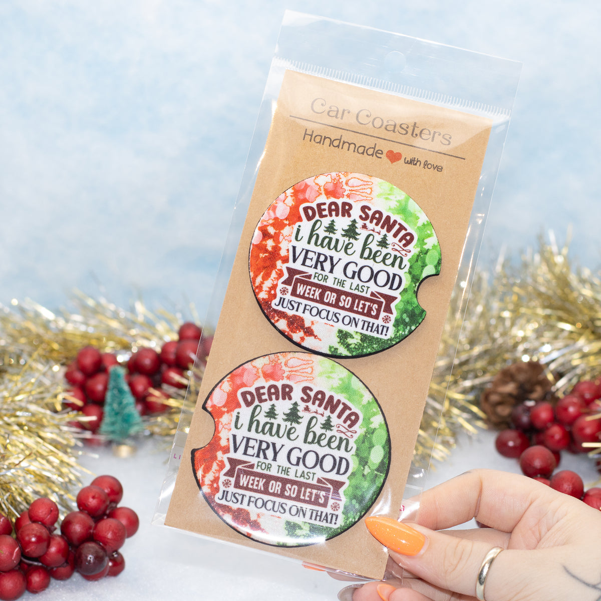 A hand two round car coasters with a holiday-themed design. The coasters feature a watercolour Christmas tree and the text 'DEAR SANTA i have been VERY GOOD FOR THE LAST WEEK ON SO LET'S JUST FOCUS ON THAT!'.