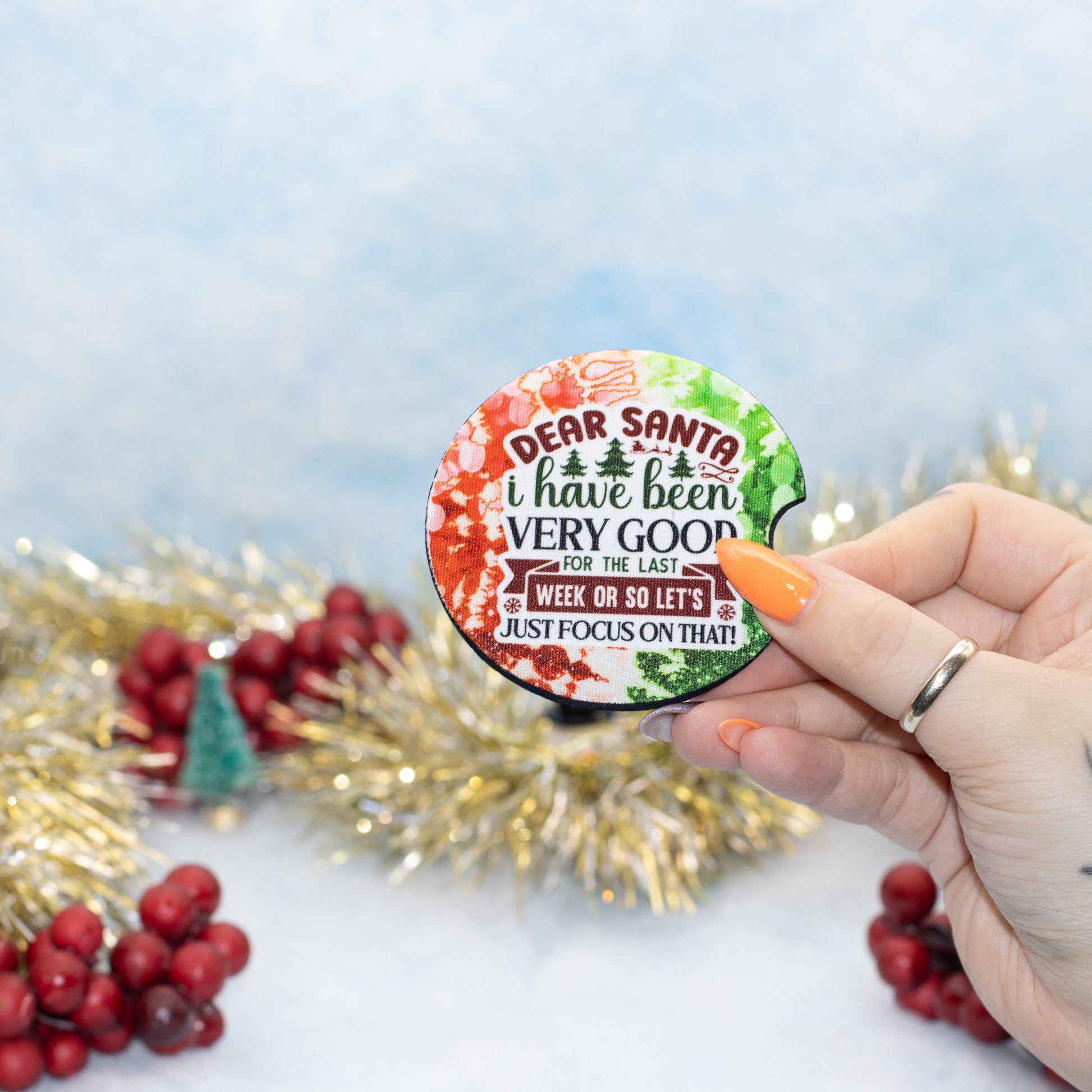 A hand holds a round car coaster that reads 'DEAR SANTA i have been VERY GOOD FOR THE LAST WEEK OR SO LET'S JUST FOCUS ON THAT!'. The pin has a festive multicolor holiday design with red, green, and white watercolor splashes and gold tinsel and red berries in the background.