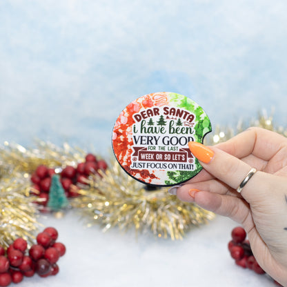 A hand holds a round car coaster that reads 'DEAR SANTA i have been VERY GOOD FOR THE LAST WEEK OR SO LET'S JUST FOCUS ON THAT!'. The pin has a festive multicolor holiday design with red, green, and white watercolor splashes and gold tinsel and red berries in the background.