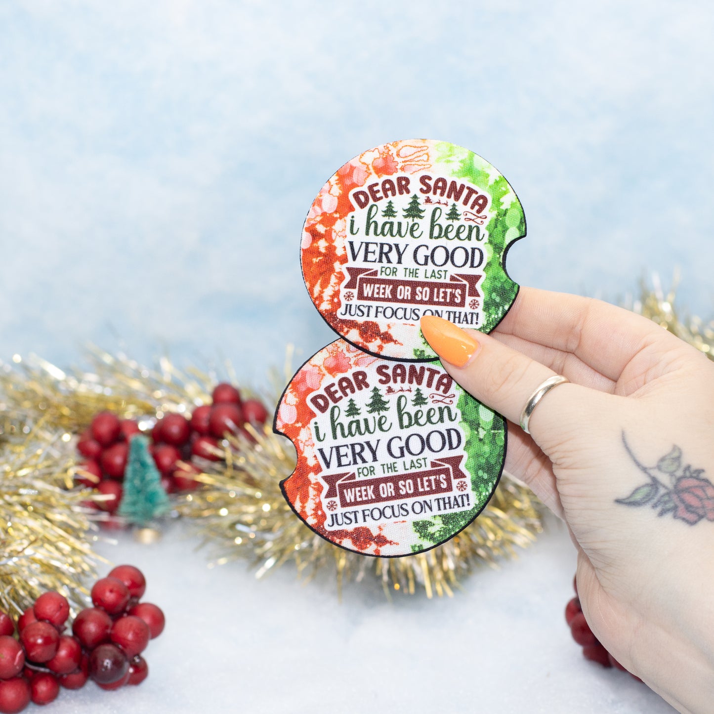 A hand holds two round Christmas-themed car coasters with the text 'DEAR SANTA I have been VERY GOOD FOR THE LAST WEEK OR SO LET'S JUST FOCUS ON THAT!', set against a festive background with gold tinsel and red berries.