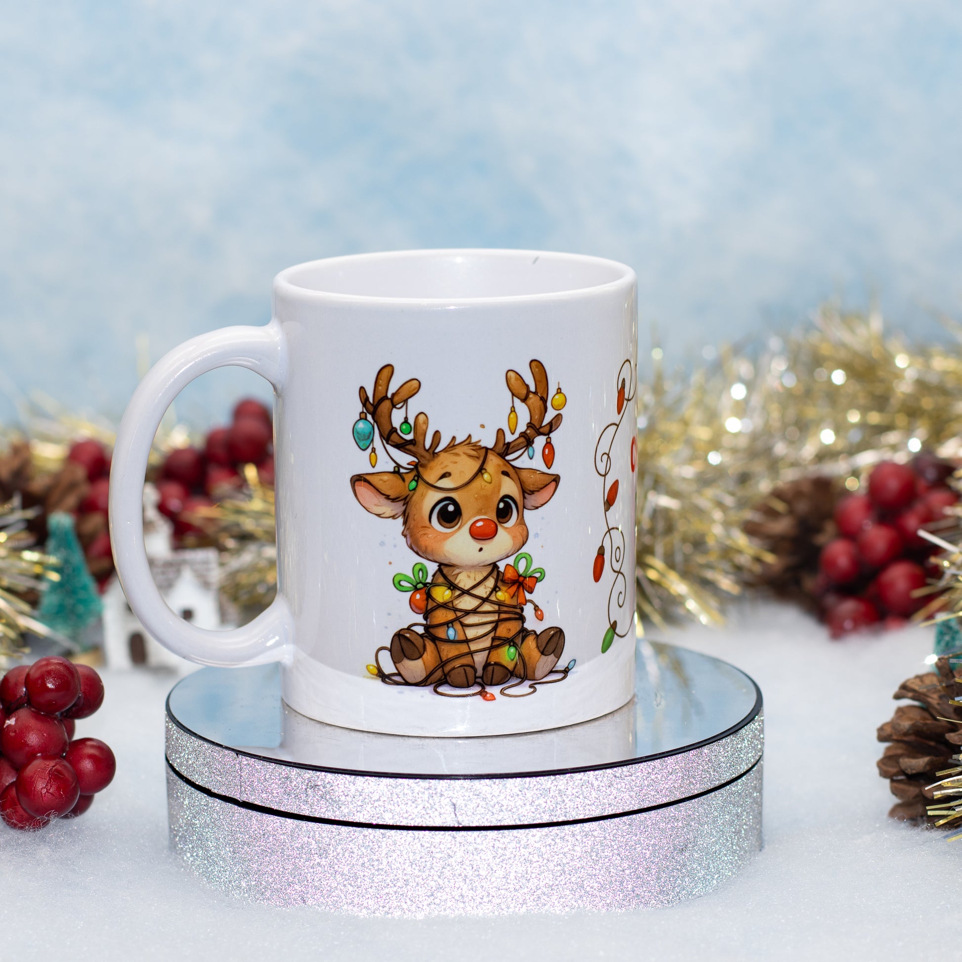 A white ceramic mug featuring a cute cartoon reindeer decorated with Christmas lights, displayed on a round silver glitter stand with festive holiday decorations in the background.