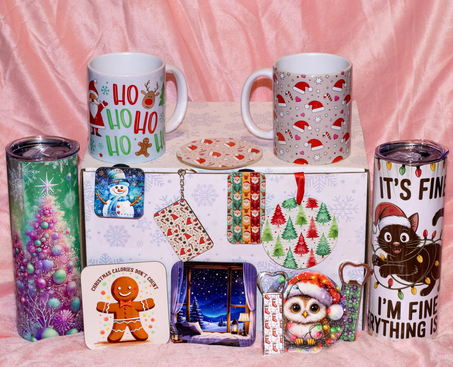 A holiday-themed gift set featuring two white mugs, two stainless steel tumblers, a coaster, and a decorative gift box with Christmas illustrations, arranged on a pink fabric background.