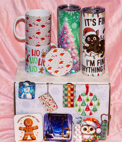 A collection of Christmas-themed mugs, tumblers, coasters, and small accessories arranged on and in front of a decorated gift box against a pink satin fabric background.