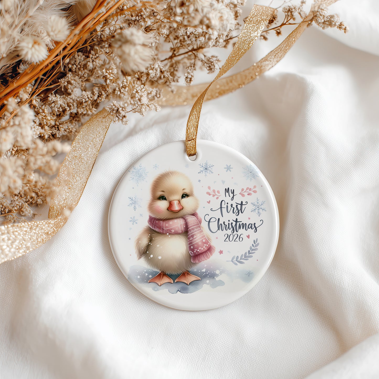 A round ceramic Christmas ornament featuring a cute cartoon duckling wearing a pink scarf, with the text 'My first Christmas 2026' and delicate snowflake and leaf illustrations. The ornament is hanging from a gold ribbon and styled on a soft white fabric with dried flowers and a gold ribbon in the background.