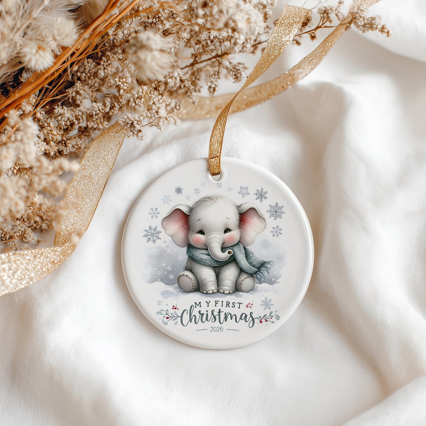 A round ceramic Christmas ornament featuring a cute cartoon baby elephant wearing a blue scarf in the snow, with the text 'MY FIRST Christmas 2026'. The ornament is styled on a soft white fabric background with a gold ribbon and dried flowers.