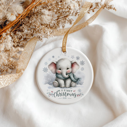 A round ceramic Christmas ornament featuring a cute cartoon baby elephant wearing a blue scarf in the snow, with the text 'MY FIRST Christmas 2026'. The ornament is styled on a soft white fabric background with a gold ribbon and dried flowers.