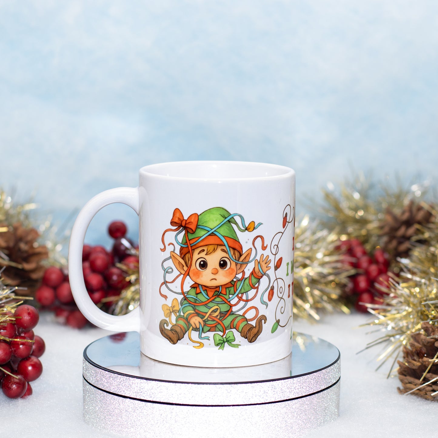 A white ceramic mug featuring a colorful cartoon elf character, displayed on a silver glittery round stand with festive holiday decorations in the background.