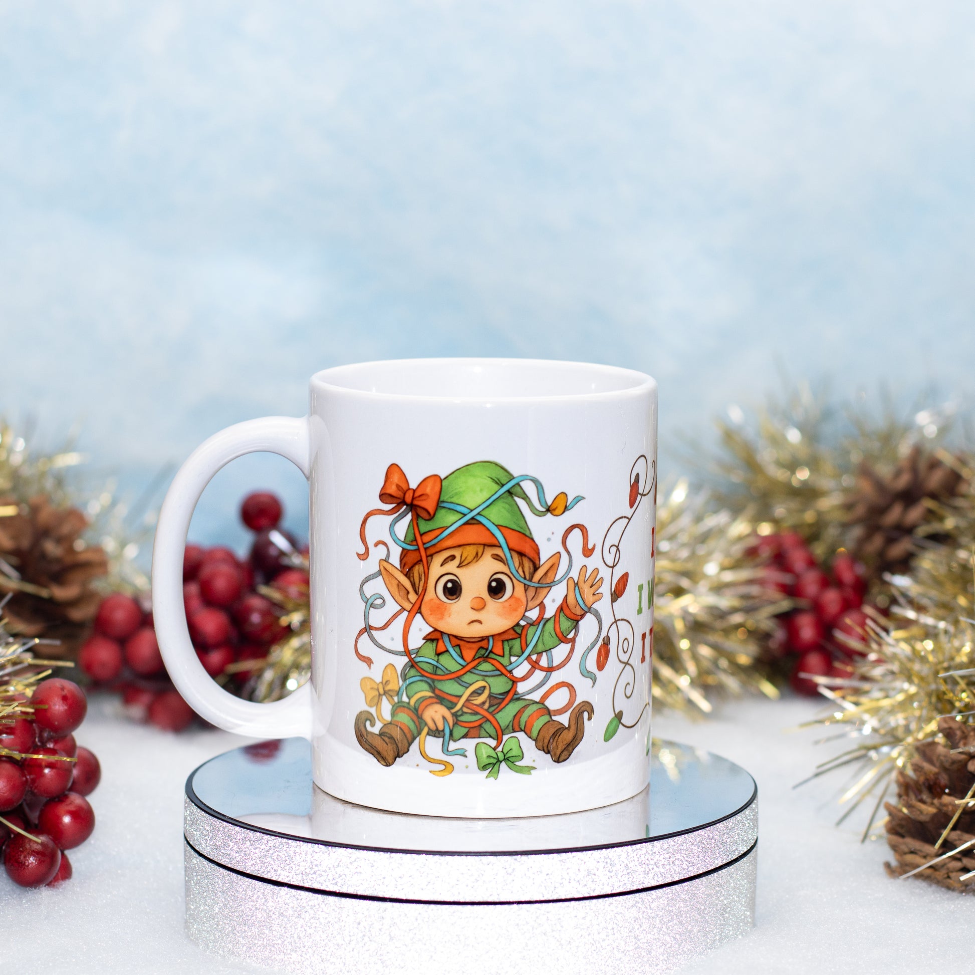 A white ceramic mug featuring a colorful cartoon elf character, displayed on a silver glittery round stand with festive holiday decorations in the background.