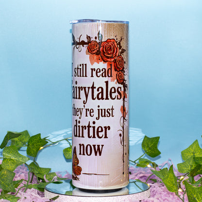 A tall insulated tumbler with a clear lid, featuring the printed phrase 'I still read fairytales they're just dirtier now' in dark brown text, decorated with orange and red roses and a butterfly on a light beige background. The tumbler is displayed on a reflective surface with decorative green ivy and pink flowers against a light blue background.
