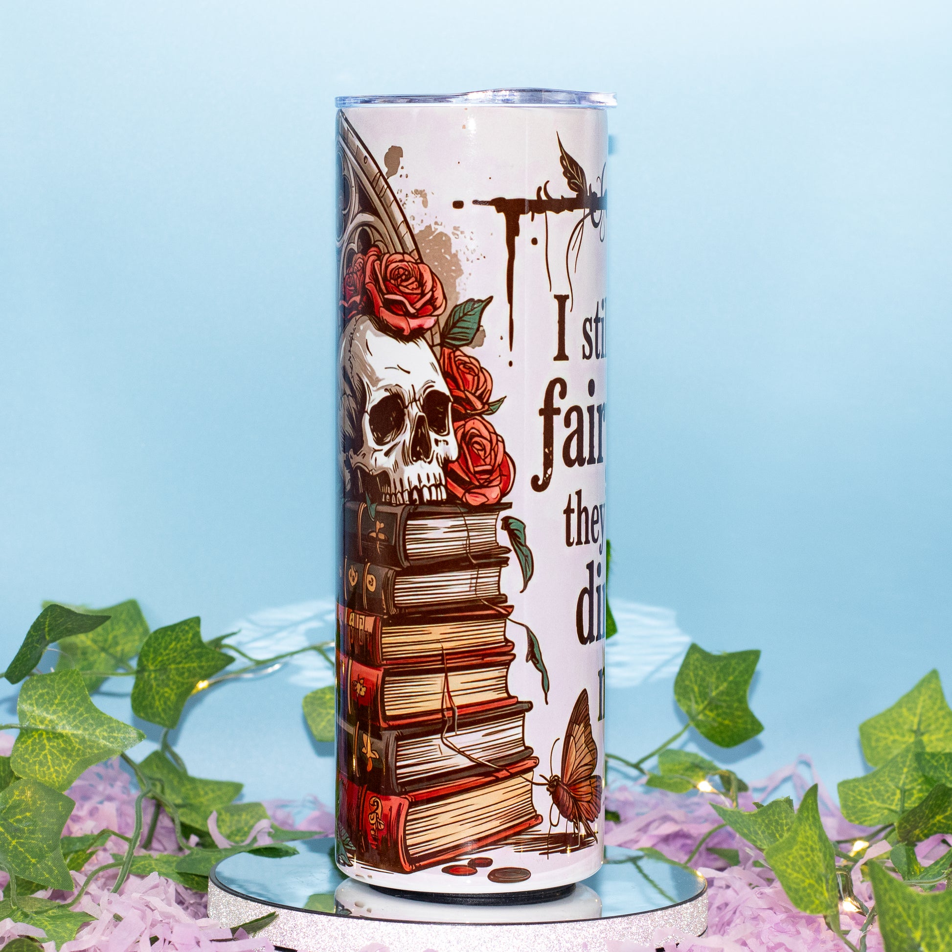 A tall insulated tumbler with a clear lid, decorated with a gothic illustration featuring a skull, stacked books, red roses, a butterfly,
