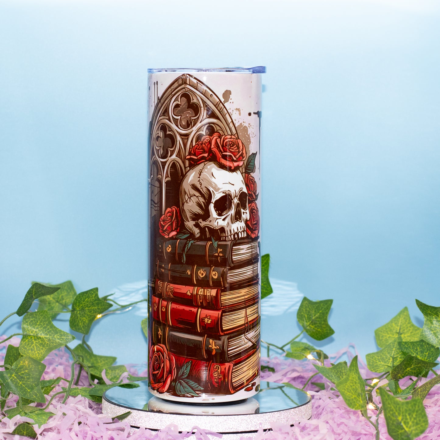 A cylindrical lamp or decorative container with a gothic illustration featuring a skull, red roses, and stacked books, displayed on a mirrored base with artificial ivy and pink petals against a light blue background.