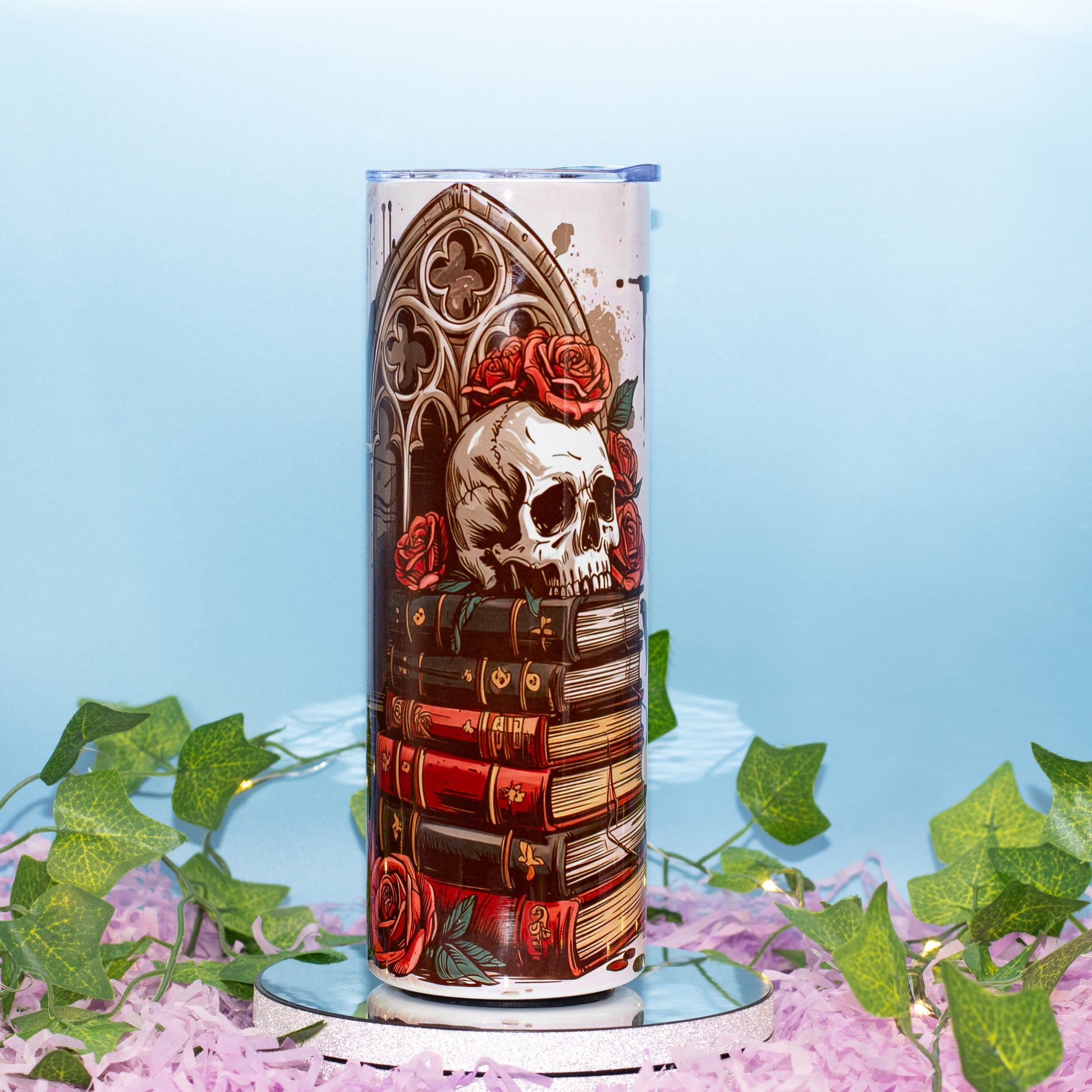 A cylindrical lamp or decorative container with a gothic illustration featuring a skull, red roses, and stacked books, displayed on a mirrored base with artificial ivy and pink petals against a light blue background.
