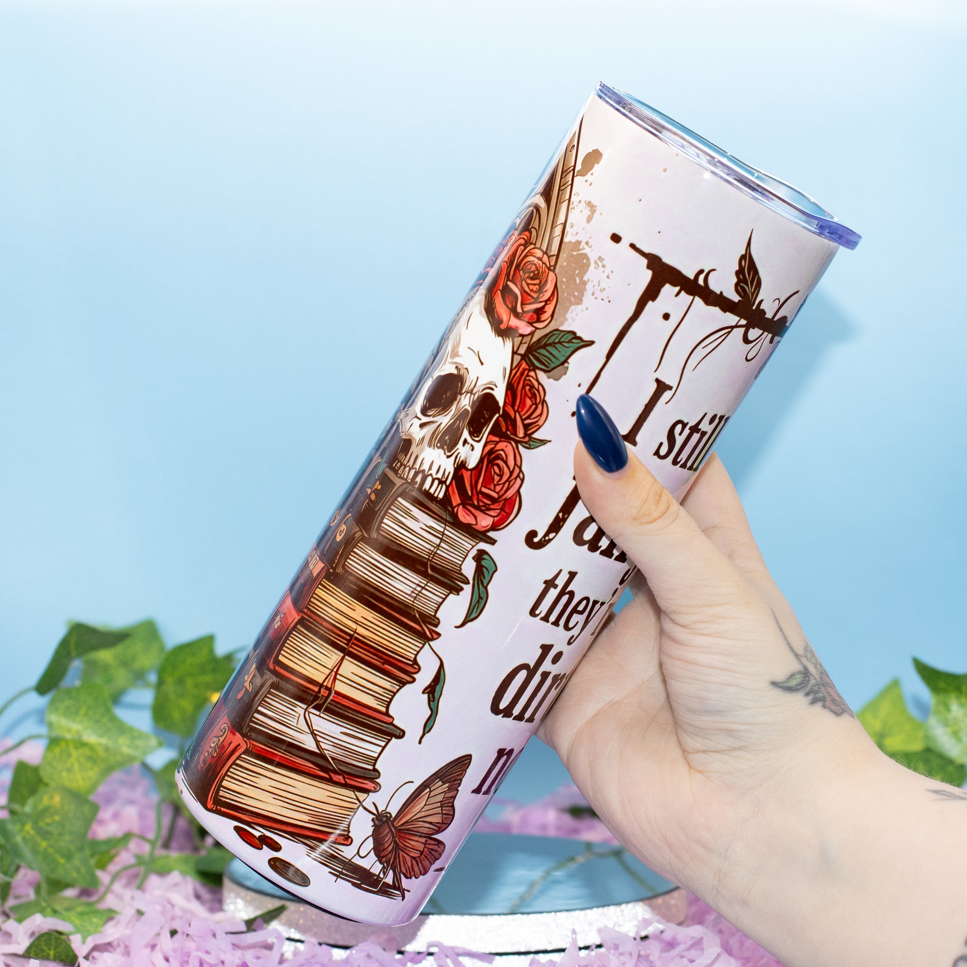 A hand holds a tall, slim tumbler with a light pink and white gothic-themed graphic featuring a skull, books, roses, and text. The background is a soft light blue with decorative green leaves and purple flowers at the bottom.