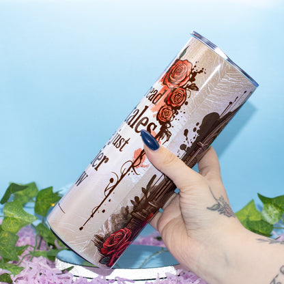 A hand holds a slim stainless steel tumbler featuring a vintage-style design with red roses, dark brown ink splatters, and partial text on a light blue background with decorative green leaves and purple flowers.