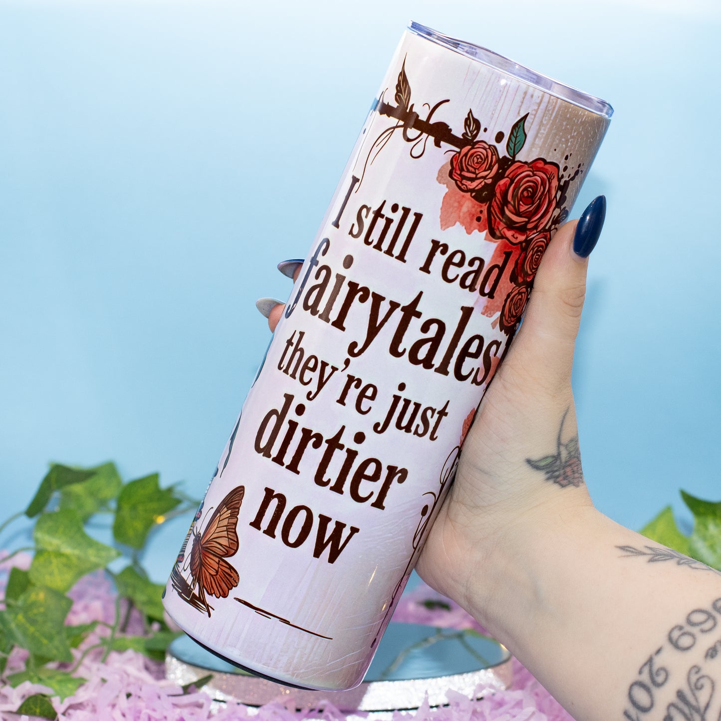 A hand holds a white insulated tumbler featuring the printed quote 'I still read fairytales they're just dirtier now' alongside red roses and brown butterflies, set against a light blue background with decorative green leaves and pink petals.
