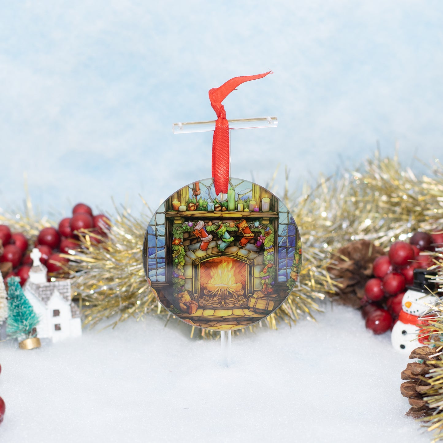 A round Christmas ornament featuring a colorful stained-glass-style fireplace scene with a red hanging ribbon, styled on a snowy surface with gold tinsel, red berries, pinecones, a small house, and a snowman.
