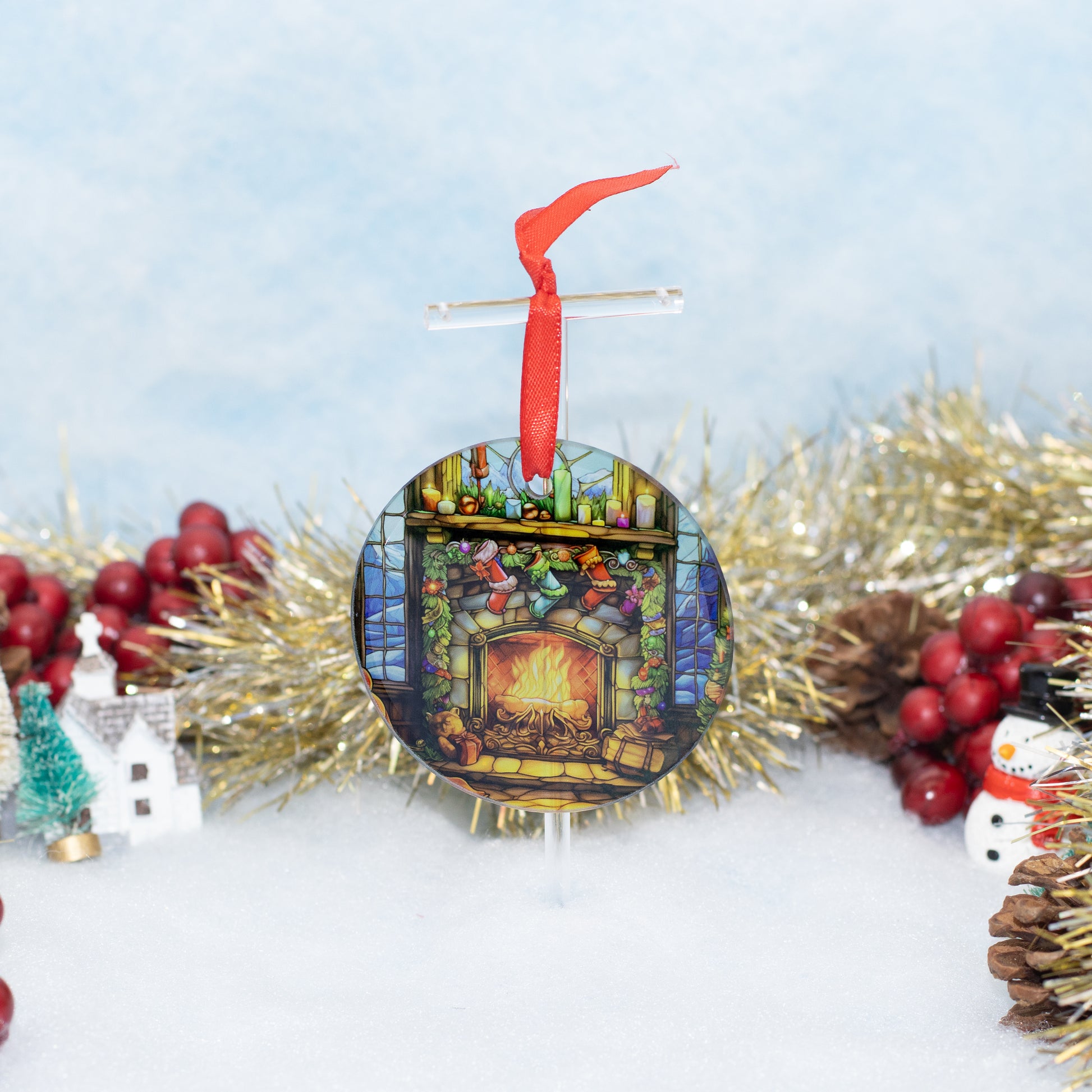 A round Christmas ornament featuring a colorful stained-glass-style fireplace scene with stockings and gifts, hanging from a red ribbon. The ornament is displayed on a stand amidst holiday decorations including gold tinsel, red berries, pinecones, a small snowman figure, and a miniature house against a soft light blue and white background.