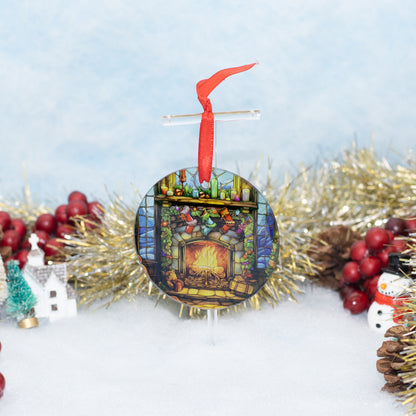 A round Christmas ornament featuring a colorful stained-glass-style fireplace scene with stockings and gifts, hanging from a red ribbon. The ornament is displayed on a stand amidst holiday decorations including gold tinsel, red berries, pinecones, a small snowman figure, and a miniature house against a soft light blue and white background.