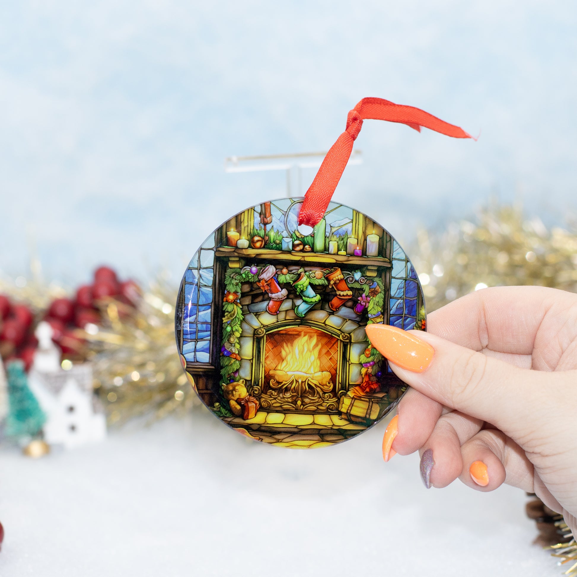 A hand holds a round Christmas ornament featuring a stained-glass-style fireplace scene with stockings, a red ribbon, and gold tinsel in the background.