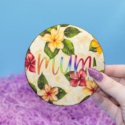A hand holds a round fabric coaster featuring a tropical floral pattern and the word 'mum' in colourful script, set against a blue background with purple shredded paper.