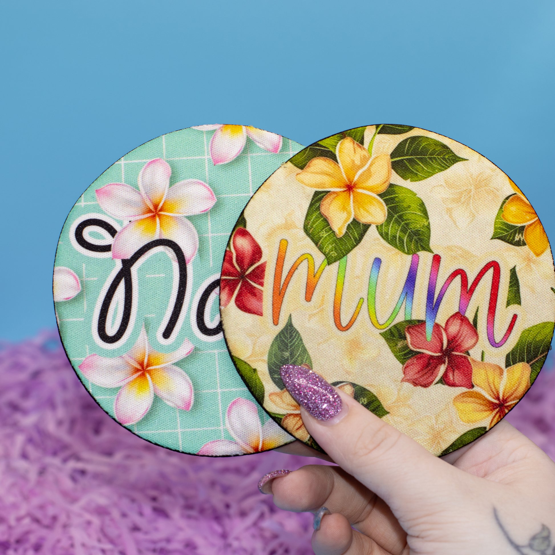 Two round coasters with tropical floral designs and the word 'Mum' in rainbow letters, held by a hand against a blue background with purple shredded paper.