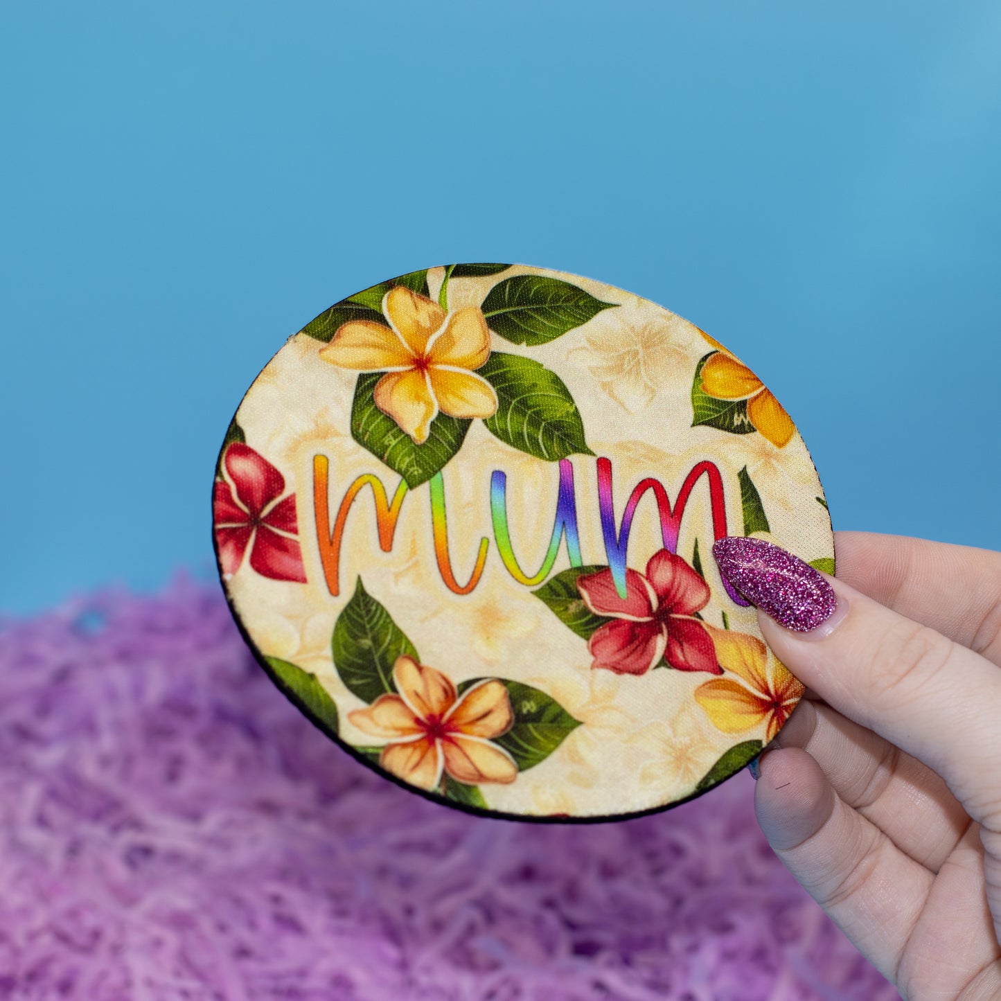 A hand holds a round wooden plaque featuring a tropical floral pattern and the word 'mum' in rainbow-coloured letters, set against a blue background with purple shredded paper.