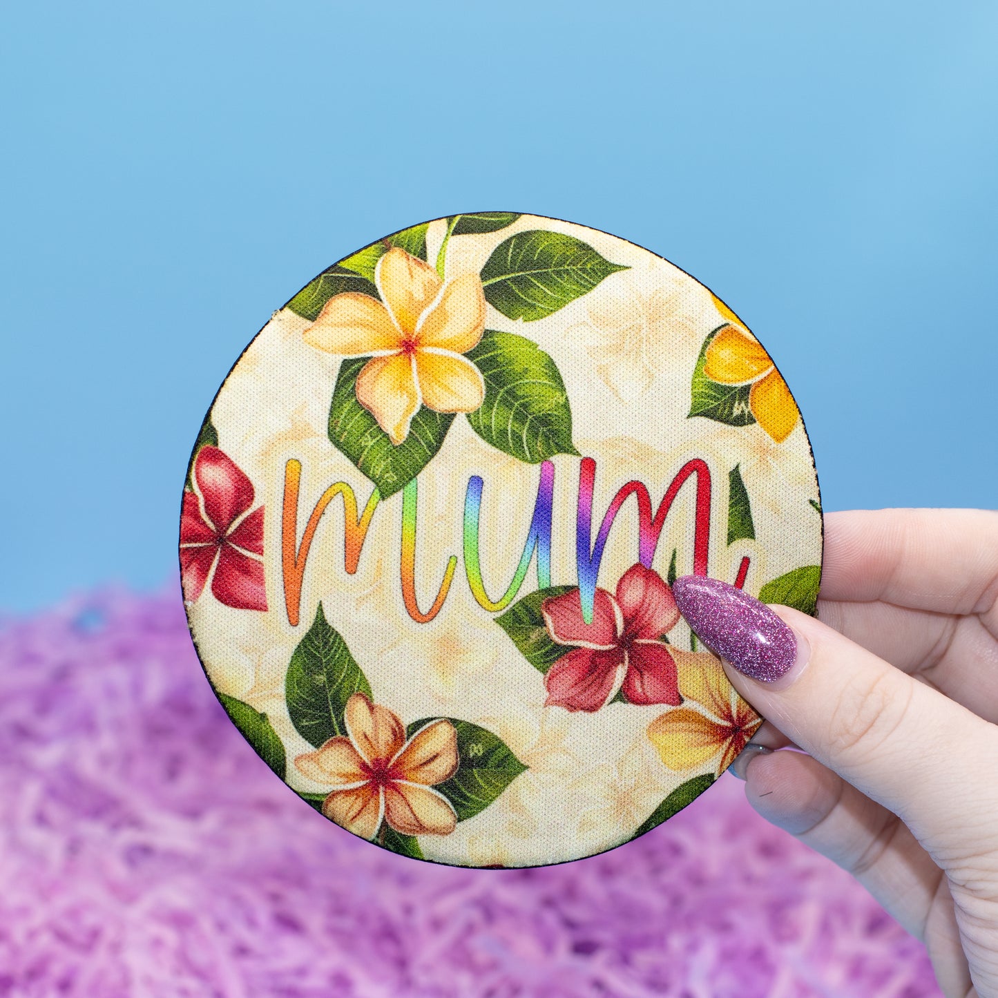 A hand holds a round coaster featuring a tropical floral design and the word 'mum' in rainbow letters, set against a blue and purple background.