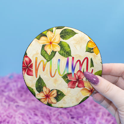 A hand holds a round coaster featuring a tropical floral design and the word 'mum' in rainbow letters, set against a blue and purple background.
