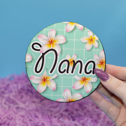 A hand holds a round custom-printed coaster featuring the text 'Nana' in black script over a mint green plumeria floral pattern, set against a blue background with purple shredded paper.