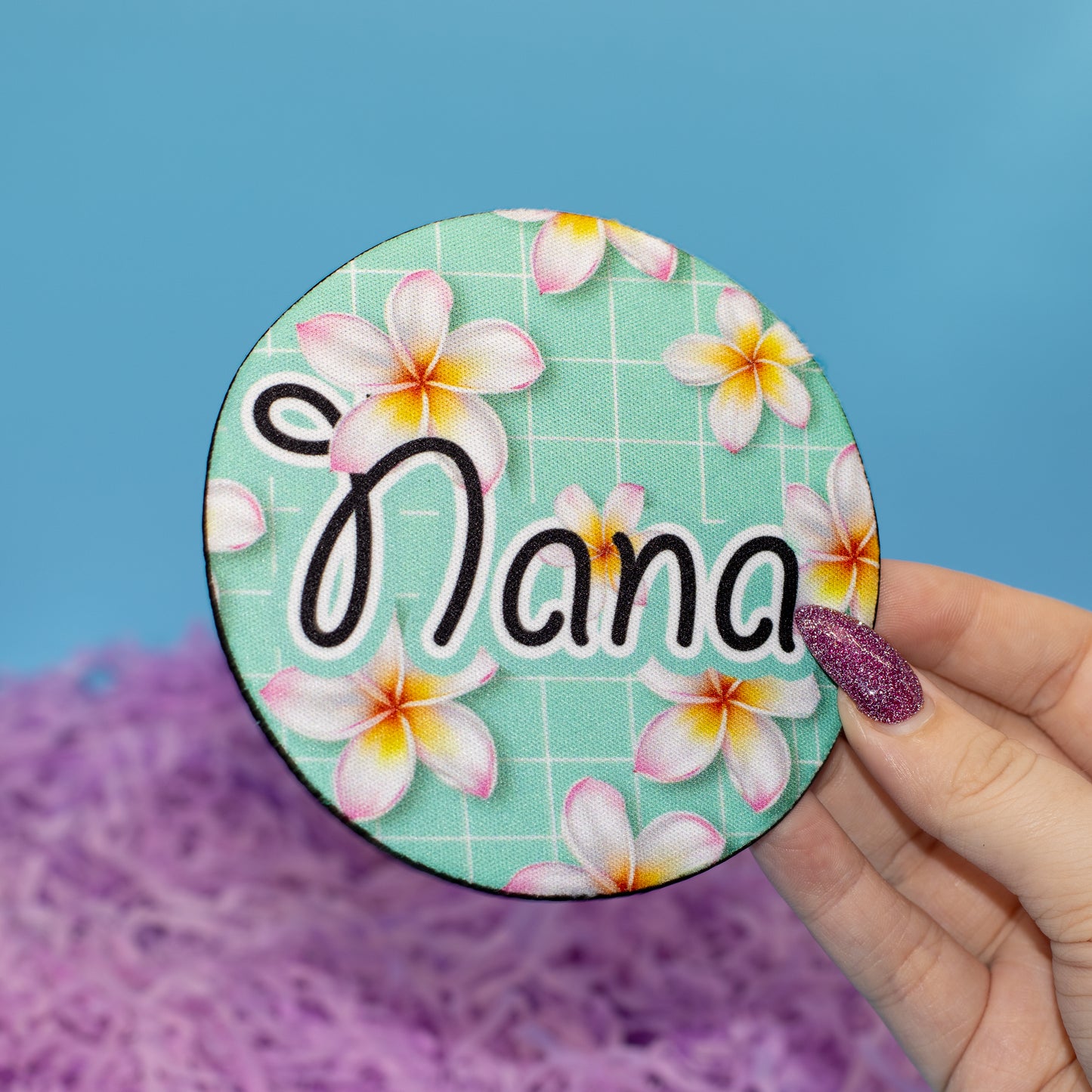 A hand holds a round embroidered patch featuring Nana in black script over a mint green plumeria floral pattern. The background consists of a solid blue wall and purple shredded paper filler.