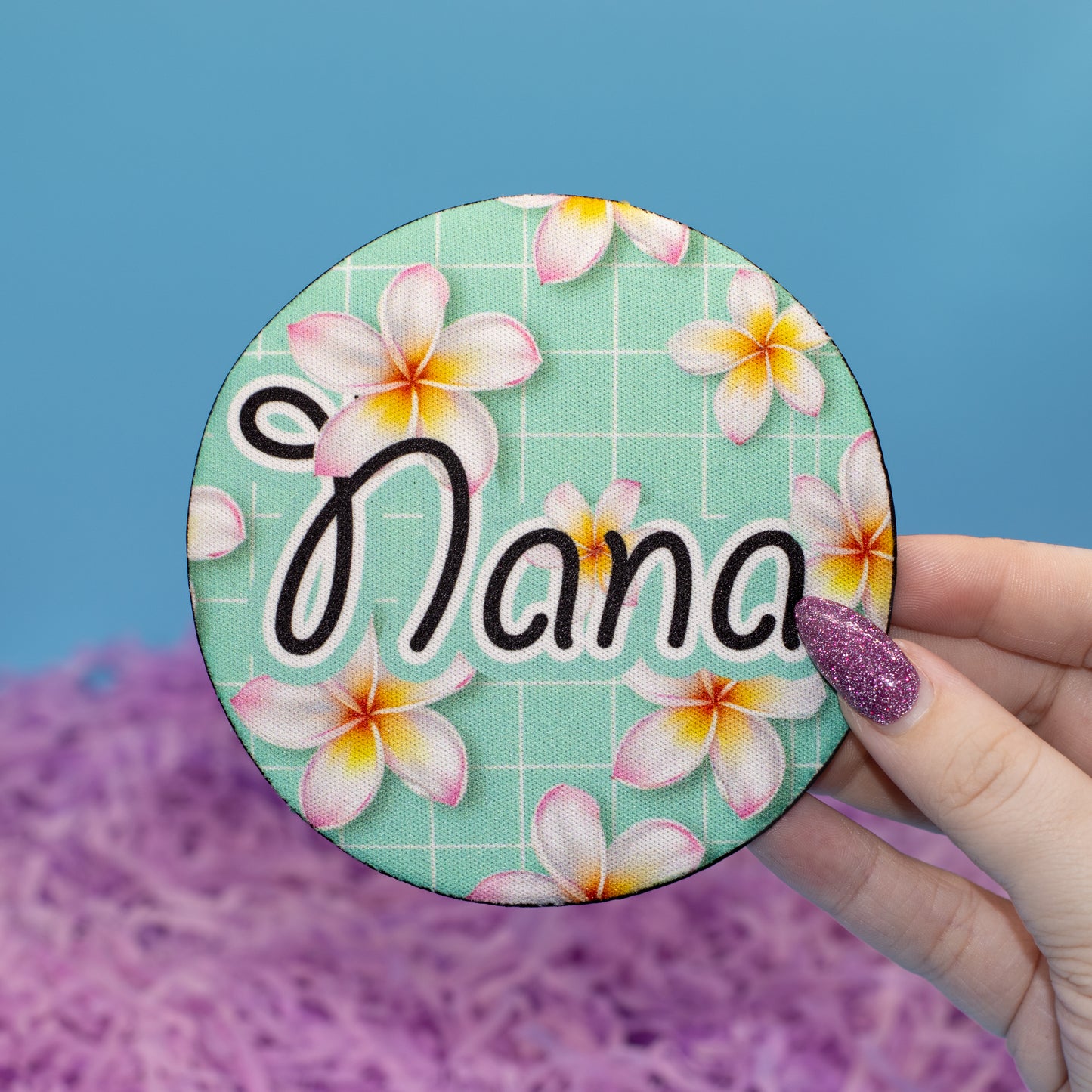 A hand holds a round coaster against a blue background with purple shredded paper. The pin features a mint green plaid pattern decorated with pink and yellow plumeria flowers and the word 'Nana' in black script.