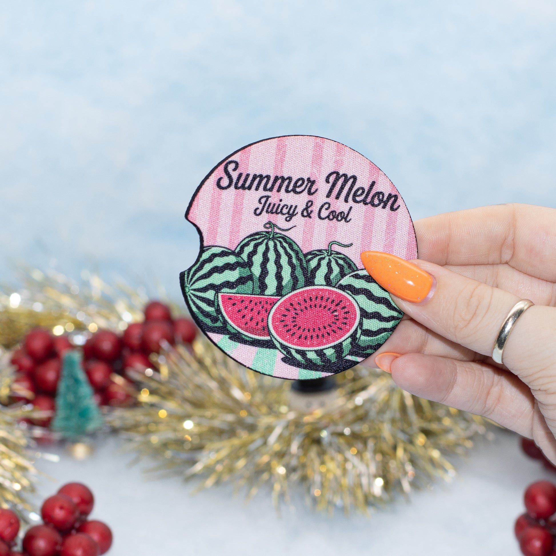 A hand holds a round car coaster featuring watermelon illustrations and the text 'Summer Melon Juicy & Cool' against a light blue background with festive tinsel and red berries.