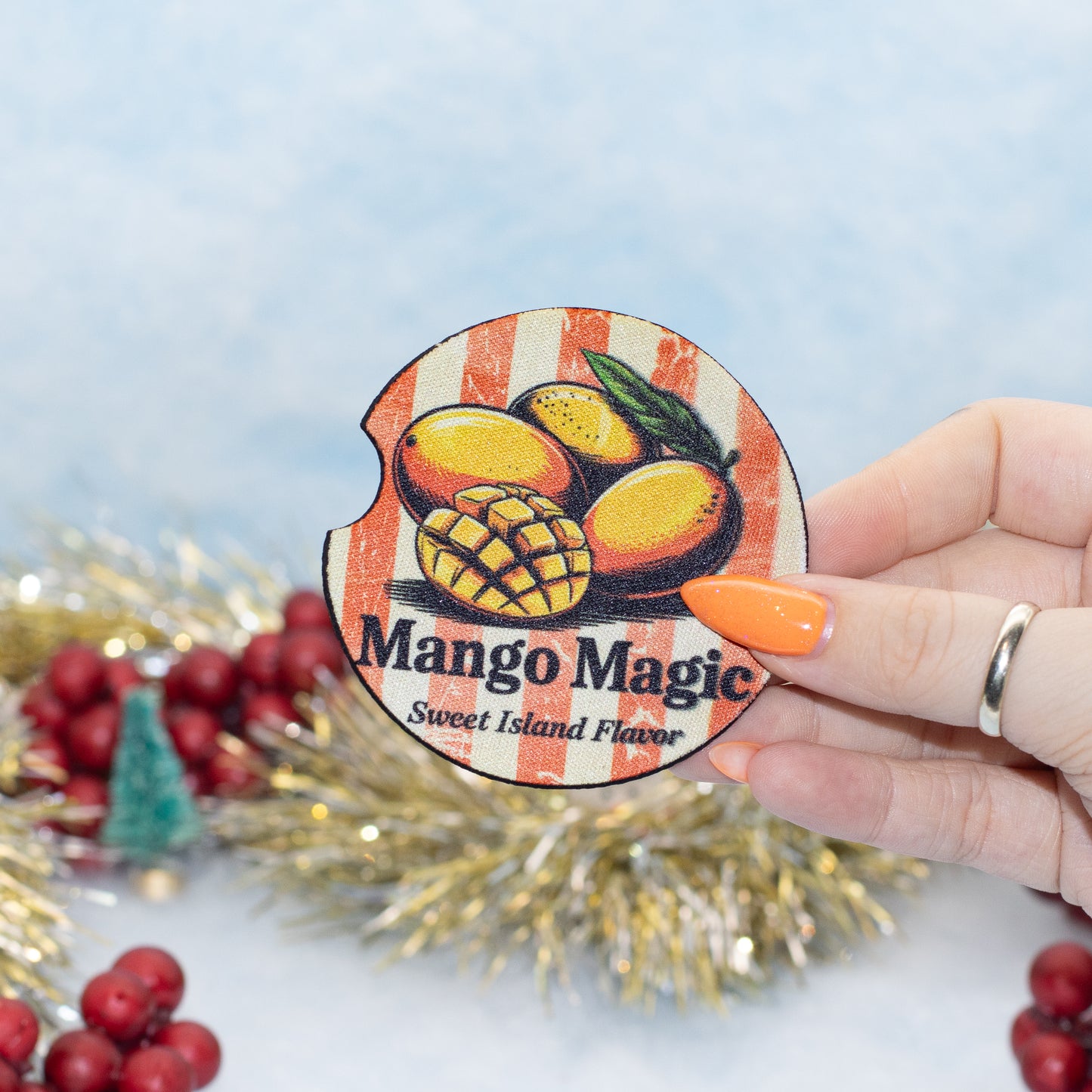 A hand holds a round car coaster that reads 'Mango Magic Sweet Island Flavor' with illustrations of mangoes. The background is decorated with gold tinsel and red berries against a light blue backdrop.