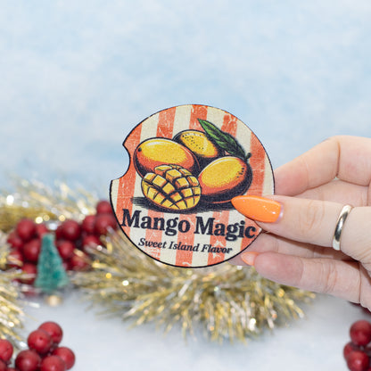 A hand holds a round car coaster that reads 'Mango Magic Sweet Island Flavor' with illustrations of mangoes. The background is decorated with gold tinsel and red berries against a light blue backdrop.