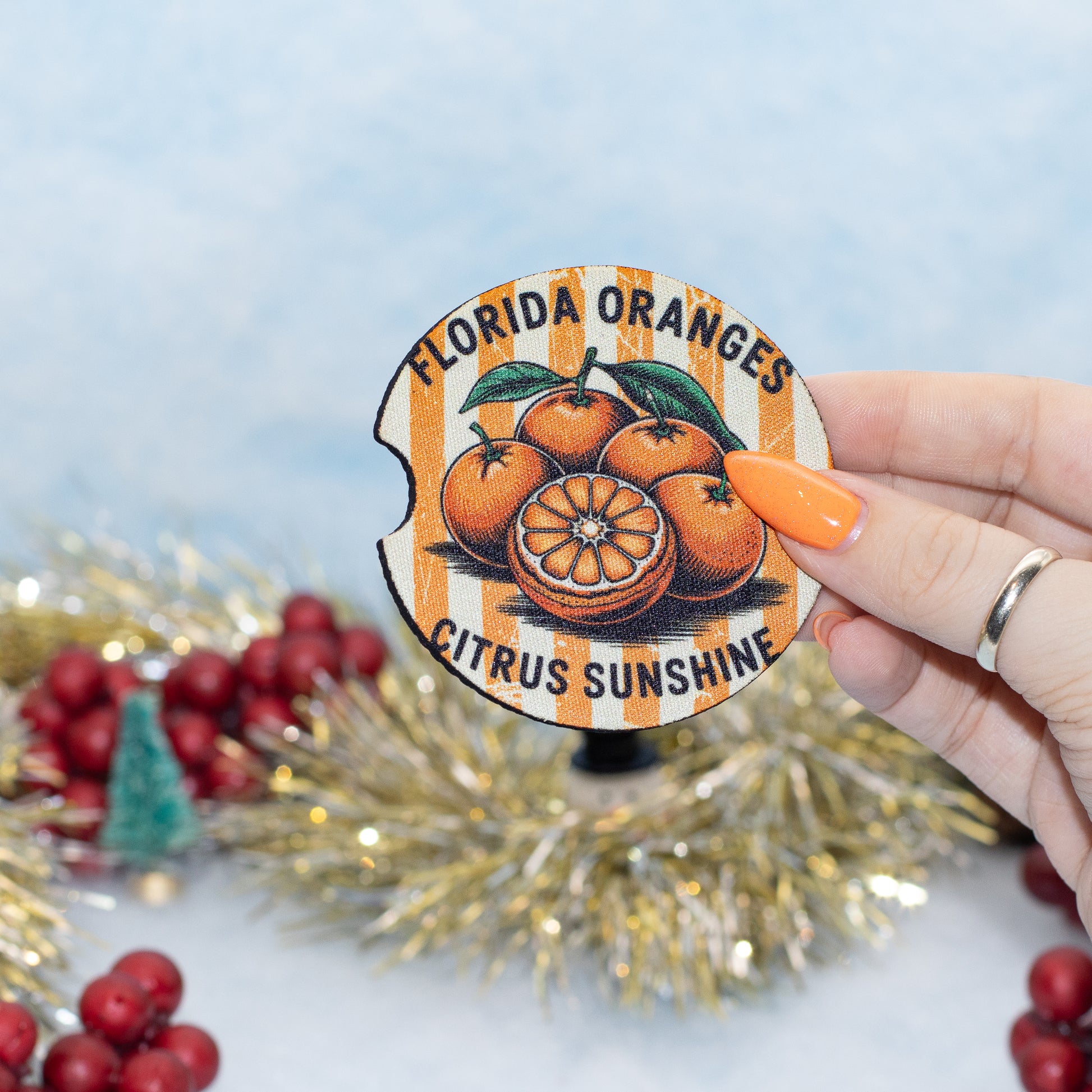 A hand holds a round vintage-style coaster featuring an illustration of Florida oranges and the text 'FLORIDA ORANGES' and 'CITRUS SUNSHINE'. The background is decorated with gold tinsel, red berries, and a soft blue backdrop.