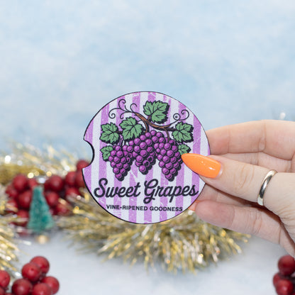 A hand holds a round, car coaster featuring a purple grape illustration and the text 'Sweet Grapes' and 'VINE-RIPENED GOODNESS' against a light blue background with festive tinsel and red berries.