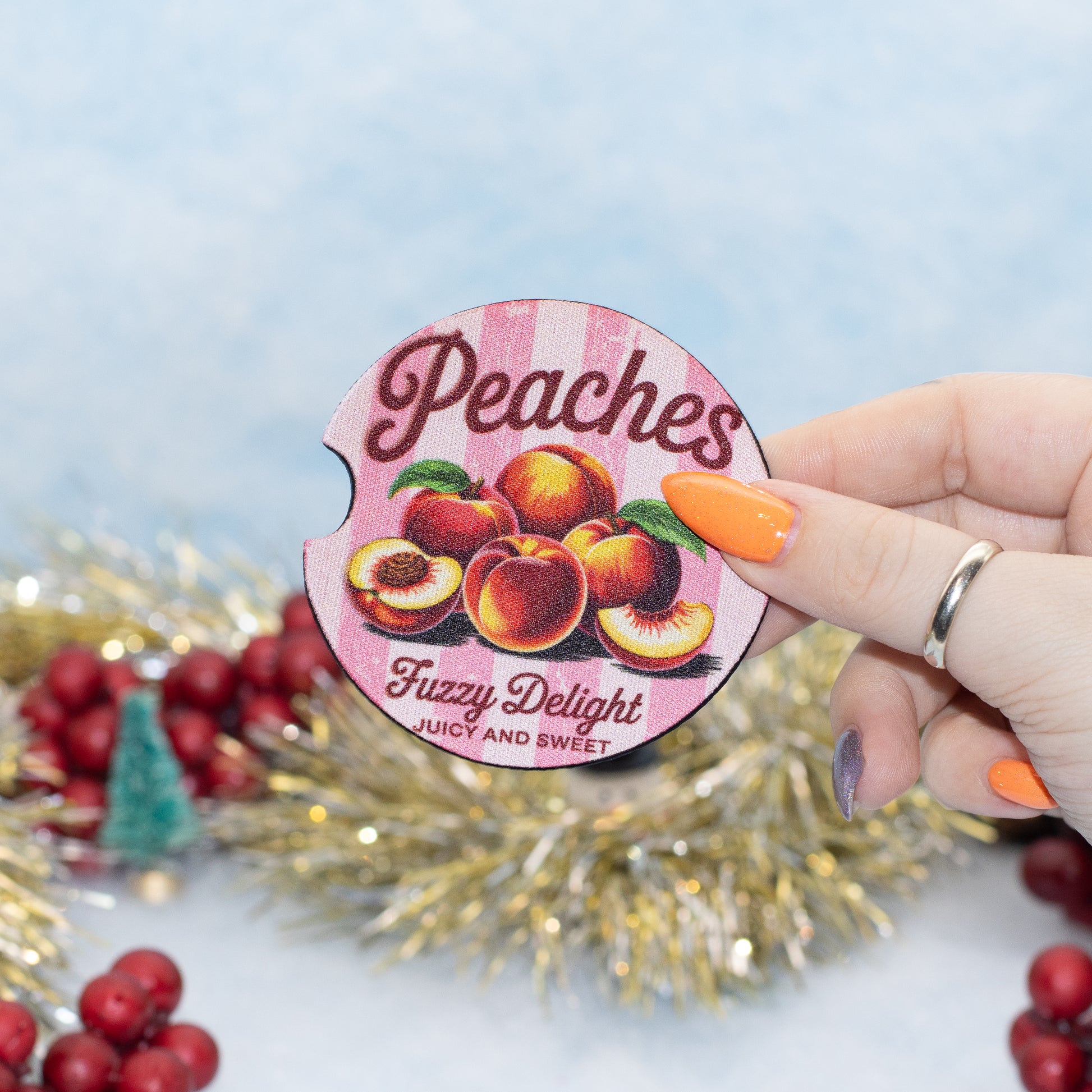 A hand holds a round, car coaster featuring a vintage-style illustration of peaches and the text 'Peaches Fuzzy Delight Juicy and Sweet' against a festive, blurred background with gold tinsel and red berries.