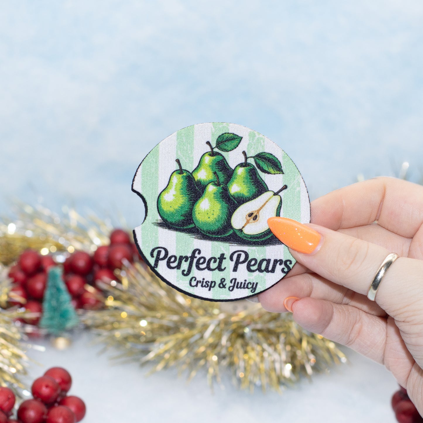 A hand holds a round car coaster featuring green pears and the text 'Perfect Pears Crisp & Juicy' against a festive holiday background with tinsel and red berries.