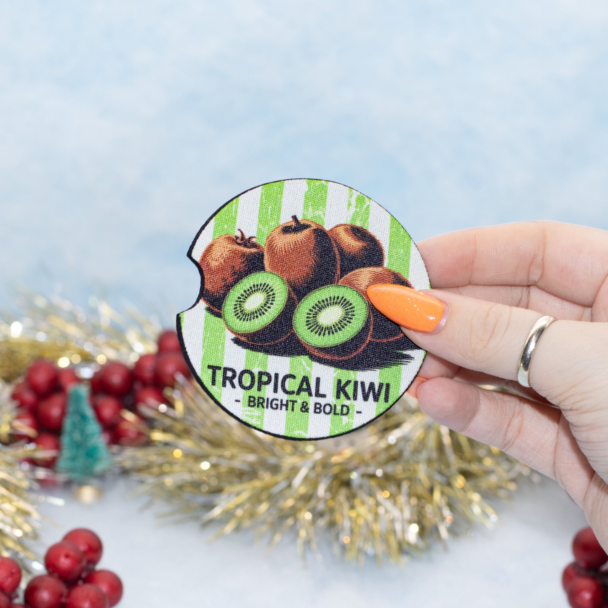 A hand holds a round car coaster featuring an illustration of tropical kiwi and apples. The sticker has green and white stripes and reads 'TROPICAL KIWI - BRIGHT & BOLD -'.