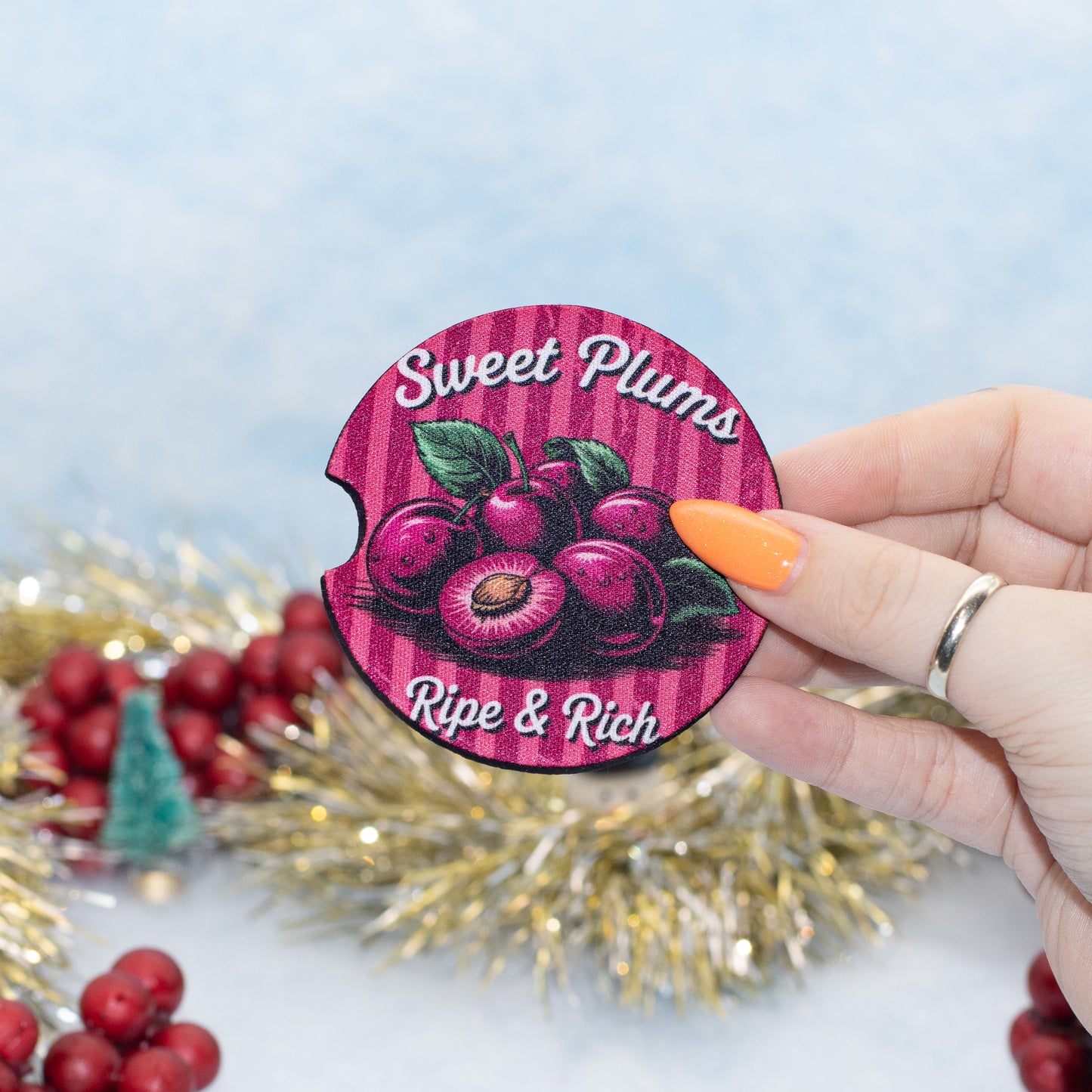 A hand holds a rounder coaster featuring a graphic of ripe sweet plums on a pink striped background with the text 'Sweet Plums' and 'Ripe & Rich'. The background is decorated with gold tinsel, red berries, and a soft blue backdrop.
