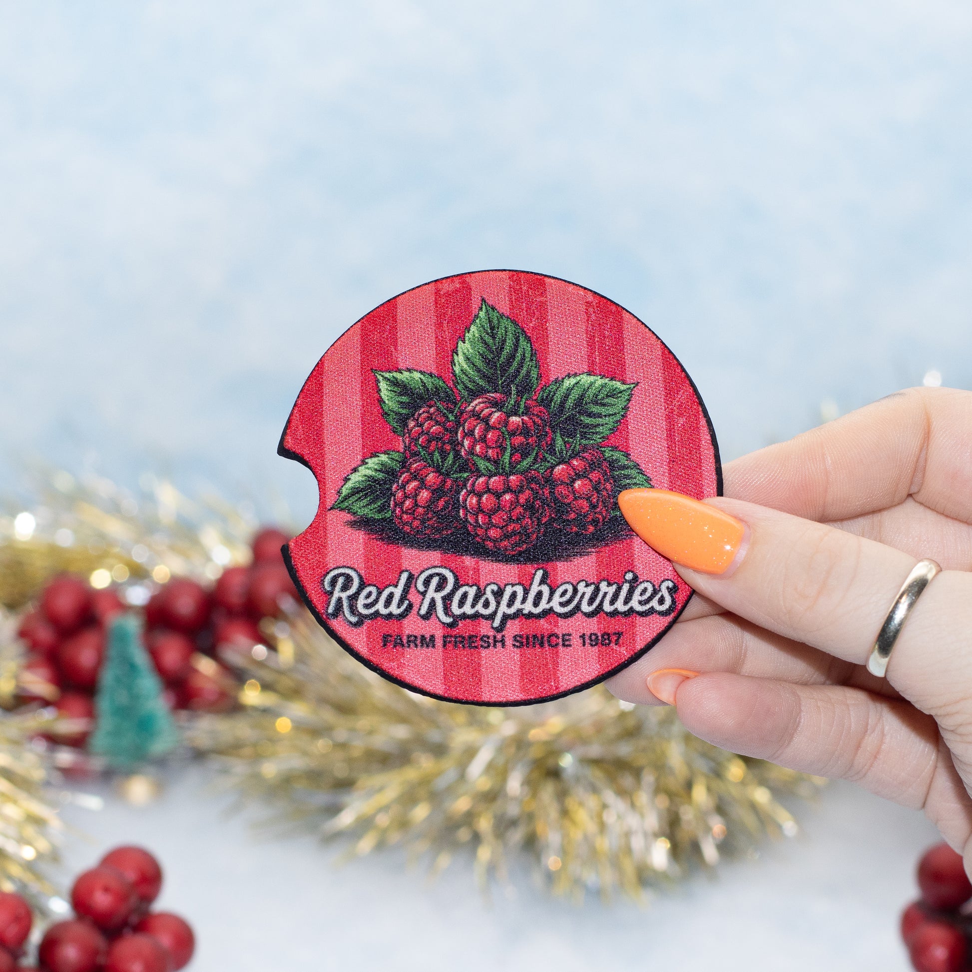 A hand holds a round red-and-pink striped car coaster featuring illustrated raspberries and the text 'Red Raspberries' and 'FARM FRESH SINCE 1987.' The background is a soft light blue with blurred holiday decorations and red berries.