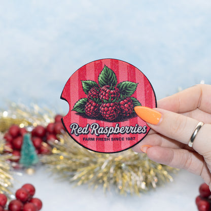 A hand holds a round red-and-pink striped car coaster featuring illustrated raspberries and the text 'Red Raspberries' and 'FARM FRESH SINCE 1987.' The background is a soft light blue with blurred holiday decorations and red berries.