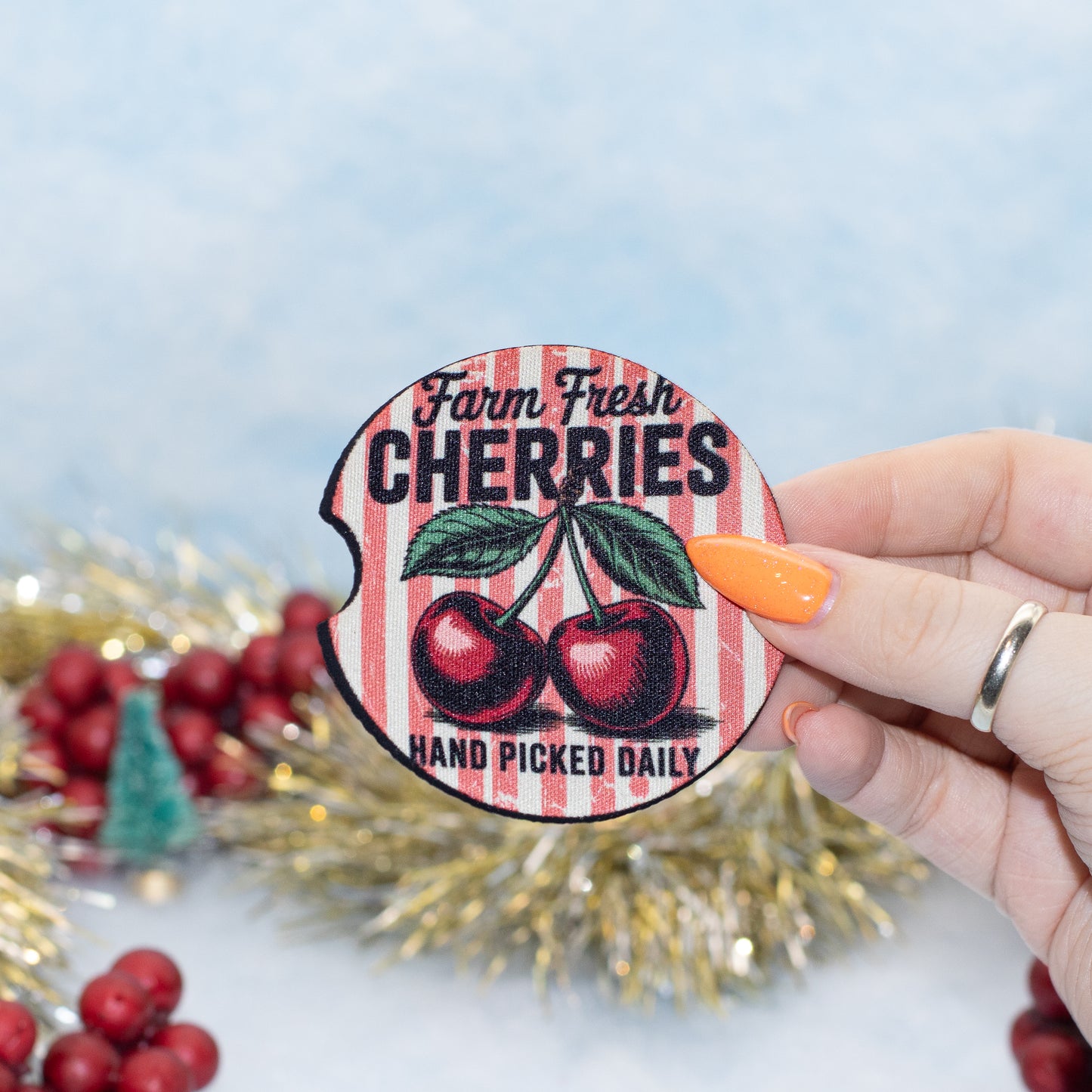 A hand holds a round car coaster that reads 'Farm Fresh CHERRIES HAND PICKED DAILY' with two red cherries on a red-and-white striped background. The scene is decorated with red berries, gold tinsel, and a soft blue backdrop.