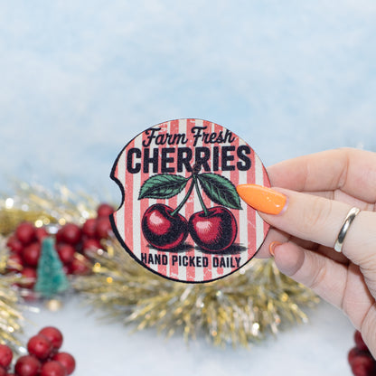 A hand holds a round car coaster that reads 'Farm Fresh CHERRIES HAND PICKED DAILY' with two red cherries on a red-and-white striped background. The scene is decorated with red berries, gold tinsel, and a soft blue backdrop.