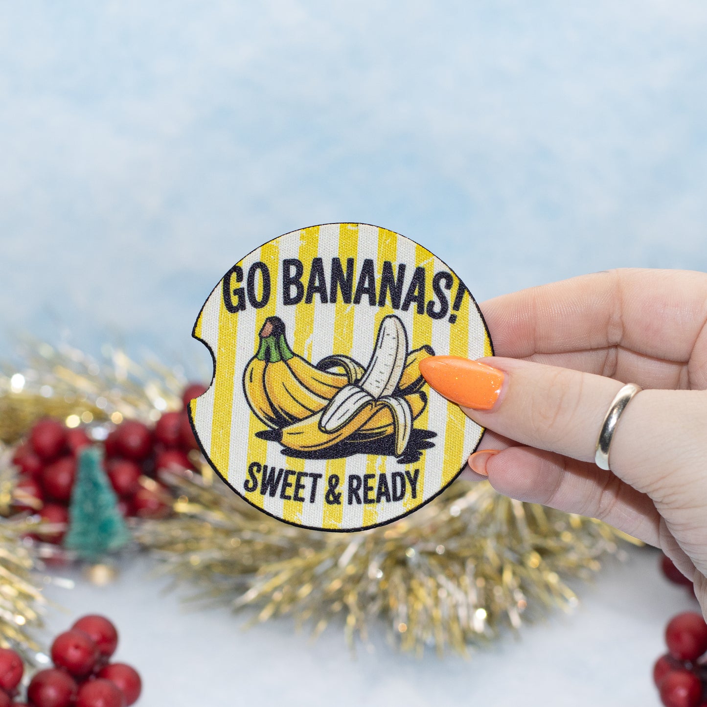 A hand holds a round car coaster featuring bananas and the text 'GO BANANAS! SWEET & READY!'. The background is decorated with gold tinsel and red berries against a light blue backdrop.