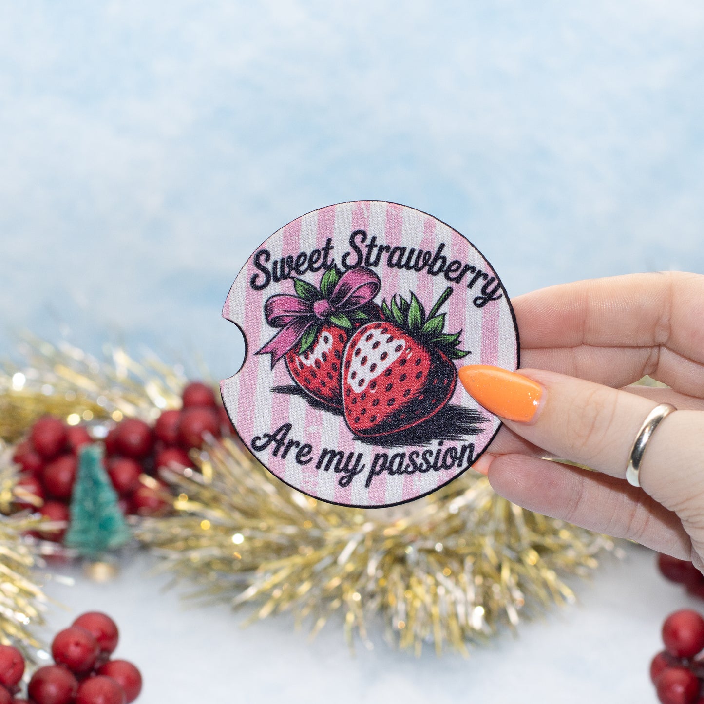 A hand holds a round car coaster featuring two strawberries and the text 'Sweet Strawberry Are my passion' against a pink striped background. The scene is decorated with gold tinsel and red berries on a light blue background.