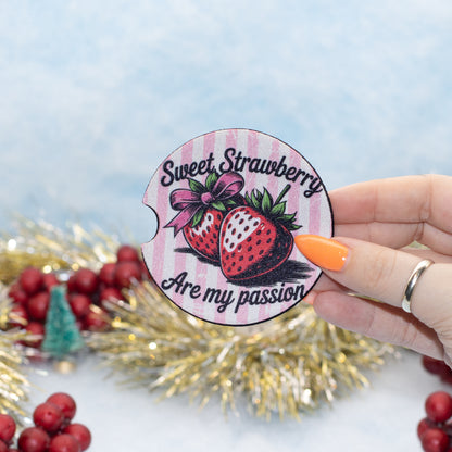 A hand holds a round car coaster featuring two strawberries and the text 'Sweet Strawberry Are my passion' against a pink striped background. The scene is decorated with gold tinsel and red berries on a light blue background.