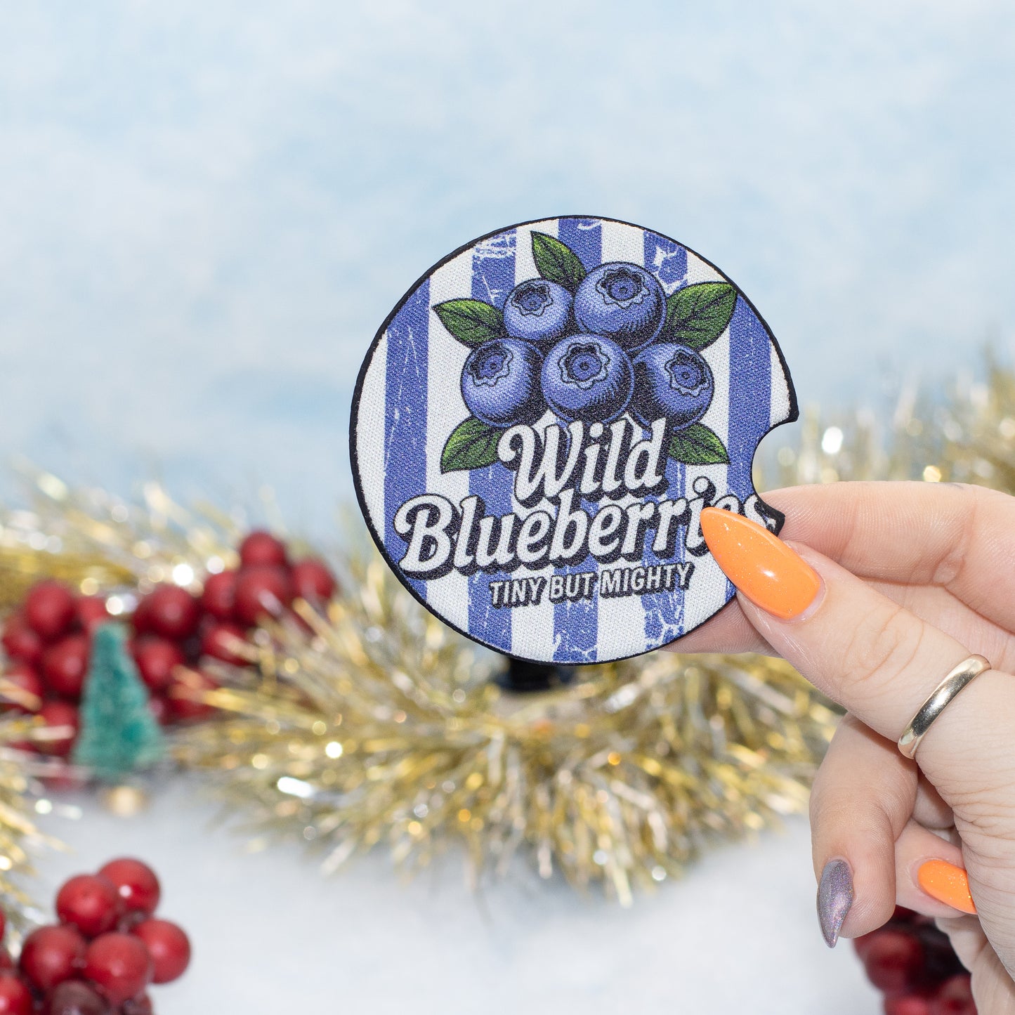 A hand holds a round blue-and-white striped coaster featuring blueberries and the text 'Wild Blueberries' and 'TINY BUT MIGHTY'. The background is a festive holiday setting with gold tinsel, red berries, and a soft light blue backdrop.