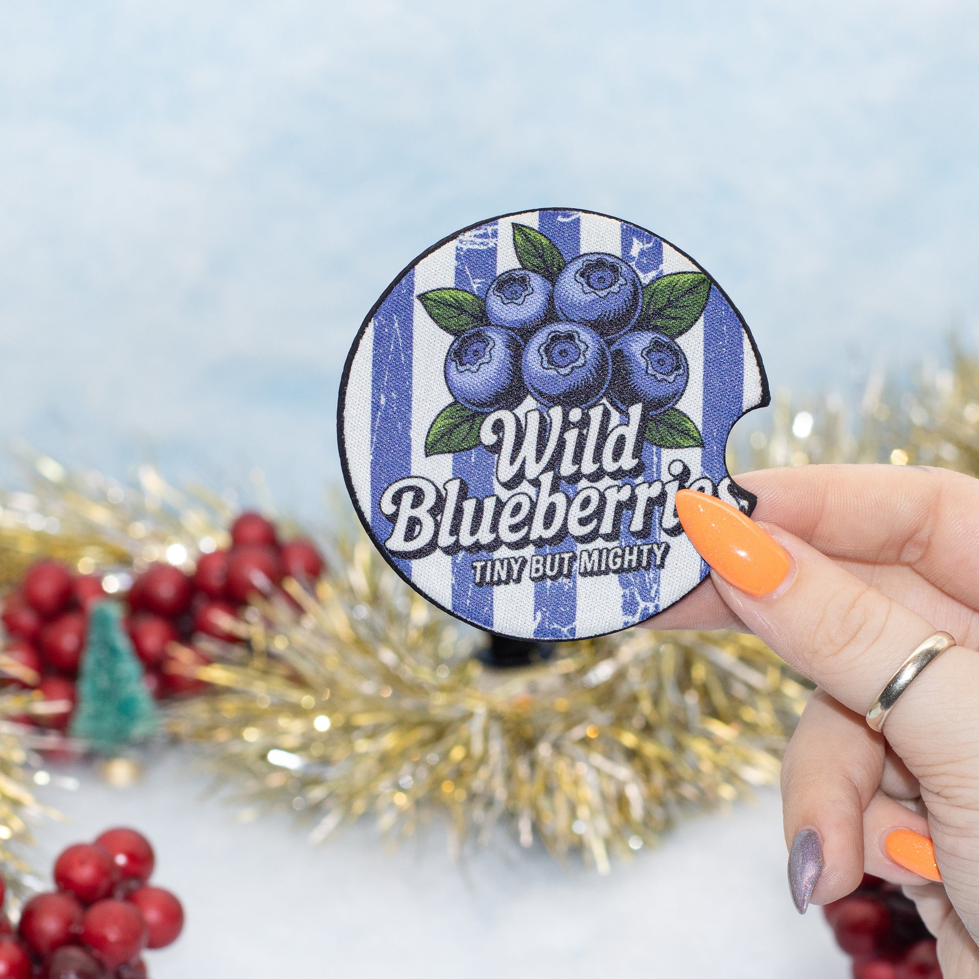 A hand holds a round blue-and-white striped coaster featuring blueberries and the text 'Wild Blueberries' and 'TINY BUT MIGHTY'. The background is a festive holiday setting with gold tinsel, red berries, and a soft light blue backdrop.